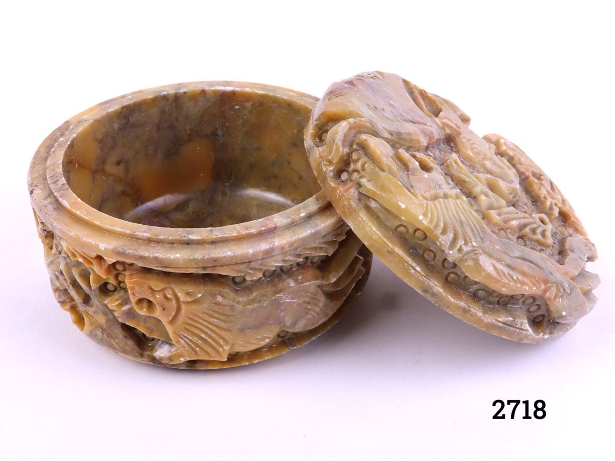 Carved soapstone lidded pot decorated with lion and elephant to the lid and lions and deer to the sides Measures 80mm in diameter Photo of pot with lid removed and leaning against pot from a near eye level angle