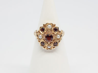 Vintage 9 karat gold ring with garnets and seed pearls. c1959 London assayed 9 karat gold ring with round cut garnet to the centre surrounded by 4 small garnets and seed pearls set on heart shaped petals with leaves on shoulders. Ring size O.5 / 7.75 Ring front measures approximately 18mm by 16mm. Main photo of ring on a display stand shown from the front