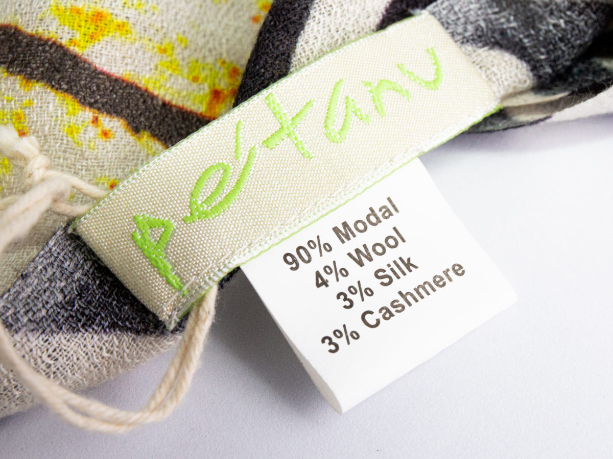 Modern hand-woven mixed fibre scarf. Luxurious hand-woven scarf in modal, wool, silk & cashmere-incredibly soft to the touch you can hardly feel it! Design is hand-printed using traditional methods of silk screen and lithographic wood block and shows pottery pieces with flowers. An absolutely gorgeous gift for someone special. Close up photo of the makers mark and list of materials on tag