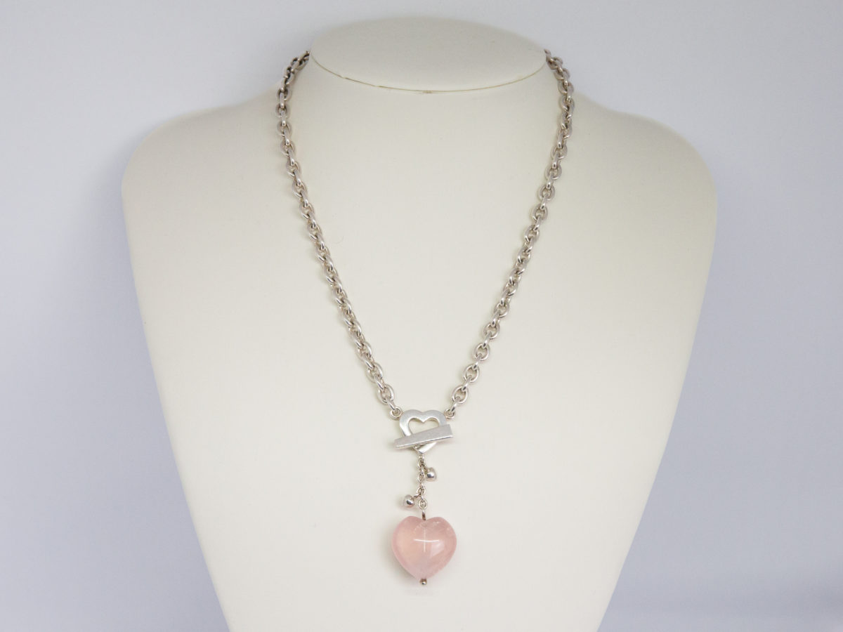 Kit Heath sterling silver and rose quartz necklace. Nice solid silver chain with an attached silver heart and a detachable rose quartz heart. The silver attachment bar is made to resemble an arrow. Rose quartz is believed to attract love and connects to the heart chakra. Detachable heart drop length is 47mm, full drop length of whole pendant area is 53mm. Hallmarked to the clasp, the attached heart and bar of detachable pendant. Main photo of necklace displayed on a stand.