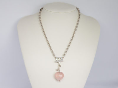 Kit Heath sterling silver and rose quartz necklace. Nice solid silver chain with an attached silver heart and a detachable rose quartz heart. The silver attachment bar is made to resemble an arrow. Rose quartz is believed to attract love and connects to the heart chakra. Detachable heart drop length is 47mm, full drop length of whole pendant area is 53mm. Hallmarked to the clasp, the attached heart and bar of detachable pendant. Main photo of necklace displayed on a stand.
