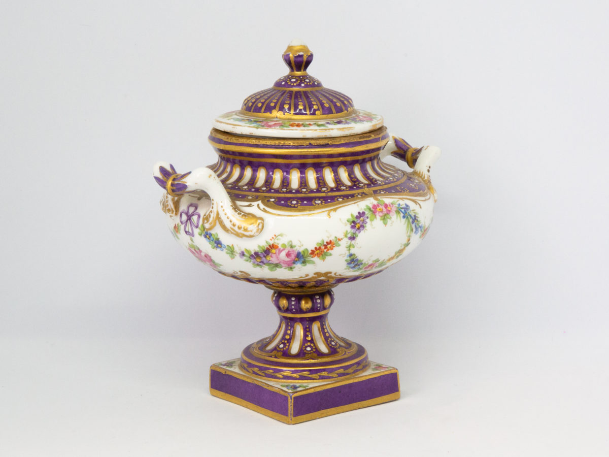 Antique Sèvres 2 handled urn with lid. Beautifully handmade and hand-painted Sèvres urn with 2 handles and lid in royal purple and gilt with floral design throughout. Some gilt wear mostly on the rim of the lid otherwise in excellent condition. Late 19th century. Base measures 55mm square, width at widest across handles 130mm, opening at top 72mm in diameter. Photo of urn shown from a side on angle with one handle to the left centre and other to top right of photo.