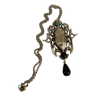 Art Nouveau style beetle necklace. Unsigned white metal necklace with an Art Nouveau style beetle pendant. The beetle is accentuated with a green bead at its head and black beads at its hind legs. A lovely statement piece. The pendant measures 85mm long and 55mm at widest point. Main photo of entire necklace with pendant to the right and chain laid to the left laid out on a flat surface.