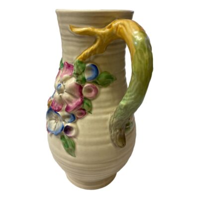 The Clarice Cliff vase is highly decorative featuring a series of colourful flowers in relief cascading down the body and a handle on one side. Photo of jug vase with the handle in the foreground at in line with the bottom right corner of the photo.