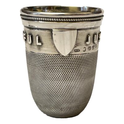 Victorian sterling silver spirit cup in the form of a thimble with raised wording at top of cup reading "Just A Thimbleful". Shield shaped empty cartouche for personalisation. c1876 Birmingham assayed by Samuel Clark. Lovely piece of historic Victorian humour? Has been well loved & used with a few dinks. Gilt interior in good condition. Measures 18mm in diameter at base and 42mm across the top. Main photo of cup seen from an eye level angle and with the empty cartouche facing centre.