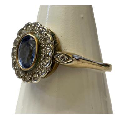 9 karat gold ring set with an oval cut lilac blue Sri Lankan sapphire within a frame of 14 round cut diamonds. Slightly larger round cut diamonds to either shoulder. Fully hallmarked for Birmingham assay c1989. Ring size Q / 8. Photo of ring on a cone shaped stand and seen from slight side angle showing the ring front and one shoulder with diamond in.
