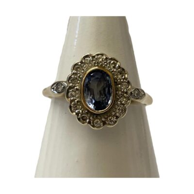9 karat gold ring set with an oval cut lilac blue Sri Lankan sapphire within a frame of 14 round cut diamonds. Slightly larger round cut diamonds to either shoulder. Fully hallmarked for Birmingham assay c1989. Ring size Q / 8. Main photo of ring on a cone shaped display stand and seen from the front.