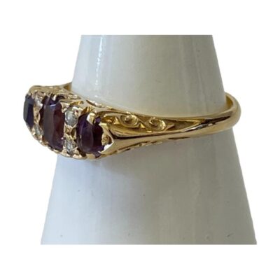 Victorian 18 karat gold trilogy ring. Pretty antique trilogy ring set with 3 oval cut amethysts which are separated with 2 round cut diamonds. The shoulders are plain in contrast to the very fancy gallery. Ring size S / 9. Photo of ring on a cone shaped stand with ring front facing towards the left and intricate detail of shoulder visible.