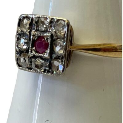 18 karat gold and platinum ring. Art Deco ring in 18 karat gold set with 8 small old mine rose cut diamonds and a 3 point ruby on a square platinum mount. Ring face measures 8mm square and weighs 2.1gms. Ring size N / 6.75. Close up photo with the ring front facing facing with part of the band on show to the right.