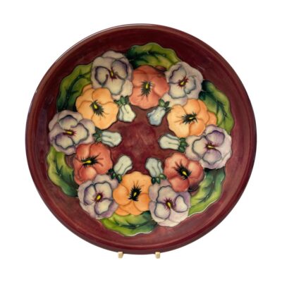 The Moorcroft Pansy plate is a beautiful example of the work of William Moorcroft. The plate has a vibrant red background and a series of colourful pansies decorating the inside. Main photo of plate seen upright and from the front.