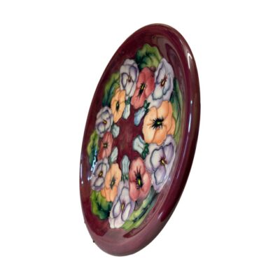 The Moorcroft Pansy plate is a beautiful example of the work of William Moorcroft. The plate has a vibrant red background and a series of colourful pansies decorating the inside. Photo of plate shown with the front facing towards the left.
