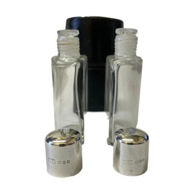 Art Deco silver topped cased bottles. A very practical cased set of 2 small half bottles with silver tops. Each bottle has sterling silver screw tops with original glass stoppers and own slot to keep secure within the case when in transit. Most likely used for cologne. Each bottle measures 53mm wide, 27mm deep at middle and 90mm tall. Full hallmark to each silver top for London assay c1926. Photo of the 2 bottles placed outside and in front of its leather case with the glass stoppers in place but silver screw tops removed and placed in the foreground with hallmarks visible on both. Seen from an eye level angle.