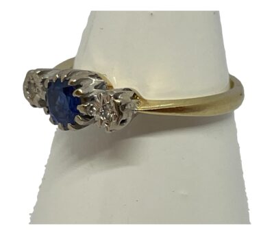 18 karat gold sapphire and diamond ring. Vintage ring in 18 karat gold with a round cut sapphire to the centre with small round cut diamonds to either side. The gem stone mount is in 18 karat white gold which accentuates the stones. Hallmarked 18ct. Ring size N.25 / 6.75. Ring weight 3.2gms Photo of ring on a cone shaped stand with ring front facing towards the left.