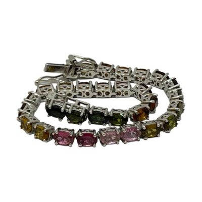Sterling silver bracelet with multi-coloured tourmaline. Modern silver tennis bracelet set with oval cut tourmaline in a variety of colours with a rainbow like look. Hallmarked 925 for sterling silver, the clasp pin end is marked metal. Main photo of bracelet laid out in a spiral on a flat surface. Some of the different colours of tourmaline are visible in the foreground and the settings for the stones seen in the background.