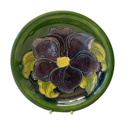 A Moorcroft shallow bowl in clematis pattern. Mid sized shallow bowl in iconic Moorcroft green with a single large purple clematis flower covering the whole bowl centre. c1947-1953. Measures 190mm across the top, 100mm at the base and 35mm deep. Main photo of plate displayed on a stand and seen forward facing with one large purple clematis flower to the centre.