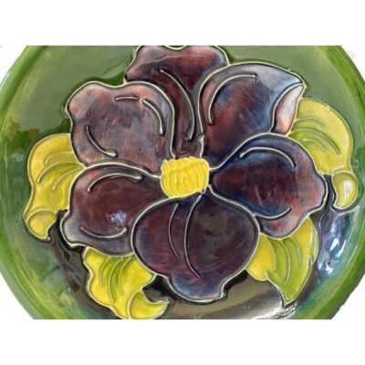 A Moorcroft shallow bowl in clematis pattern. Mid sized shallow bowl in iconic Moorcroft green with a single large purple clematis flower covering the whole bowl centre. c1947-1953. Measures 190mm across the top, 100mm at the base and 35mm deep. Close up photo of the clematis flower which covers the whole of the inside of the bowl.