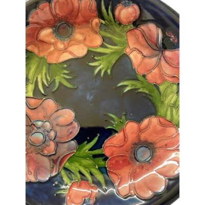 Moorcroft dish in anemone pattern. Mid sized Moorcroft dish in dark blue with light purple/red anemone flowers. Signed to the back for Walter Moorcroft and dates to between 1947-1953. Impressed stamps to the base and painted initials. Measures 110mm at base and 220mm across the top. Close up photo of the anemone flowers that decorate the inside of dish.