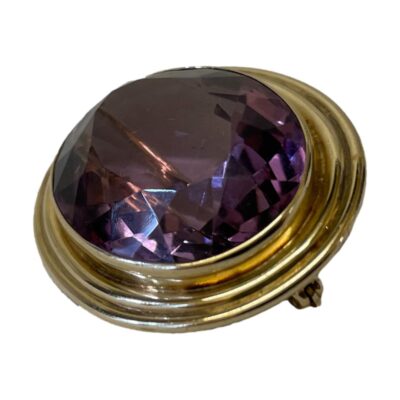 14 karat gold brooch with amethyst. Elegant 14 karat gold brooch with a large faceted amethyst to the centre. The brooch features a double security clasp with the pin shaped to lie tightly across the stone. There is also a rectangular hook for wearing as a pendant. Photo of brooch laid on a flat surface and take from a diagonal side angle with the brooch front facing upward to the top left.