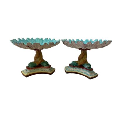 Pair of Art Nouveau sweetmeat dishes. Colourful pair of Art Nouveau majolica sweetmeat dishes. Each triple footed dish has 3 dolphins entwined to create the column which houses a scallop shaped shell like dish. The colour combination of turquoises and sea greens against pinks and yellow give these pieces are true under the sea look. Some chips to the paint and some scallop edges. Each dish measures approximately 105mm by 105mm at the base, 103mm tall and 143mm in diameter to the dish at top. Photo of the dishes side by side with the turquoise of the inside of bowl peeking through.
