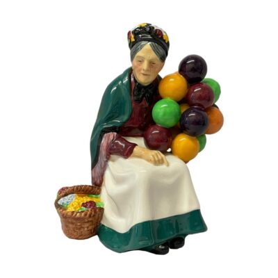 The old balloon seller by Royal Doulton. Bone china figurine of The Old Balloon Seller Number number H.N.1315 In excellent condition with no chips, cracks or crazing. A fine collectors piece. Main photo of figurine seen from a front eye level. The basket of flowers to her left and the balloons are held to the right. Her spare hand rests on her lap.