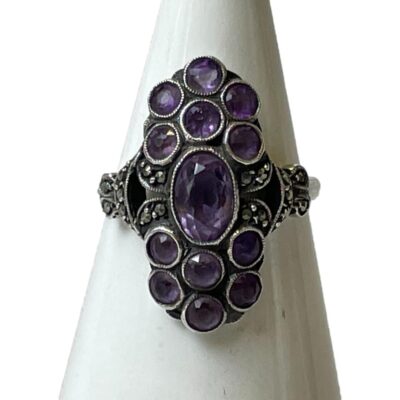 935 argentium silver ring set with amethysts. An unusual vintage ring with oval cut amethyst to the centre and a cluster of 6 round cut amethysts above and below. Accentuated with marcasite to the sides and shoulders. Very Art Deco look about it. Hallmarked 935 to inside band. Ring size N / 6.75. Main photo of ring on a cone shaped display stand with ring front facing forward. The ring front is in an oblong sort of form with the largest amethyst in oval cut at the centre.