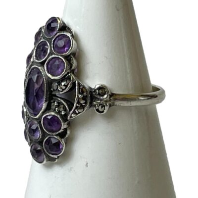 935 argentium silver ring set with amethysts. An unusual vintage ring with oval cut amethyst to the centre and a cluster of 6 round cut amethysts above and below. Accentuated with marcasite to the sides and shoulders. Very Art Deco look about it. Hallmarked 935 to inside band. Ring size N / 6.75. Photo of ring on a cone shaped stand and seen with ring front facing left.