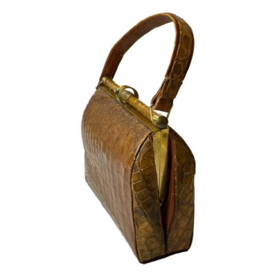1940s French crocodile skin handbag. A stylish vintage French handbag in crocodile skin with a clever shut and clip brass clasp and a small inner pocket to either side. In a well loved condition with some signs of wear. Still very useable and fashionable! Drop length from top of handle 330mm. Photo of bag seen from the side at a slight diagonal angle with width to the right foreground and one side facing towards the bottom left. The handle is extended upward.