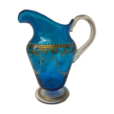 Antique Bohemian glass jug. Pretty vibrant blue jug with hand painted gilt and floral decoration. Clear glass handle and base. Late 19th century. Base measures 95mm in diameter, 185mm from spout to handle, 110mm wide and 205mm at tallest. Main photo looking at the jug from an eye level side angle with the spout to the left and handle at right. The jug is made with the spout higher than the handle.