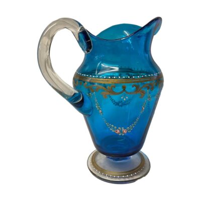 Antique Bohemian glass jug. Pretty vibrant blue jug with hand painted gilt and floral decoration. Clear glass handle and base. Late 19th century. Base measures 95mm in diameter, 185mm from spout to handle, 110mm wide and 205mm at tallest. Photo of jug seen with spout facing the top right corner and handle in the foreground to the left.