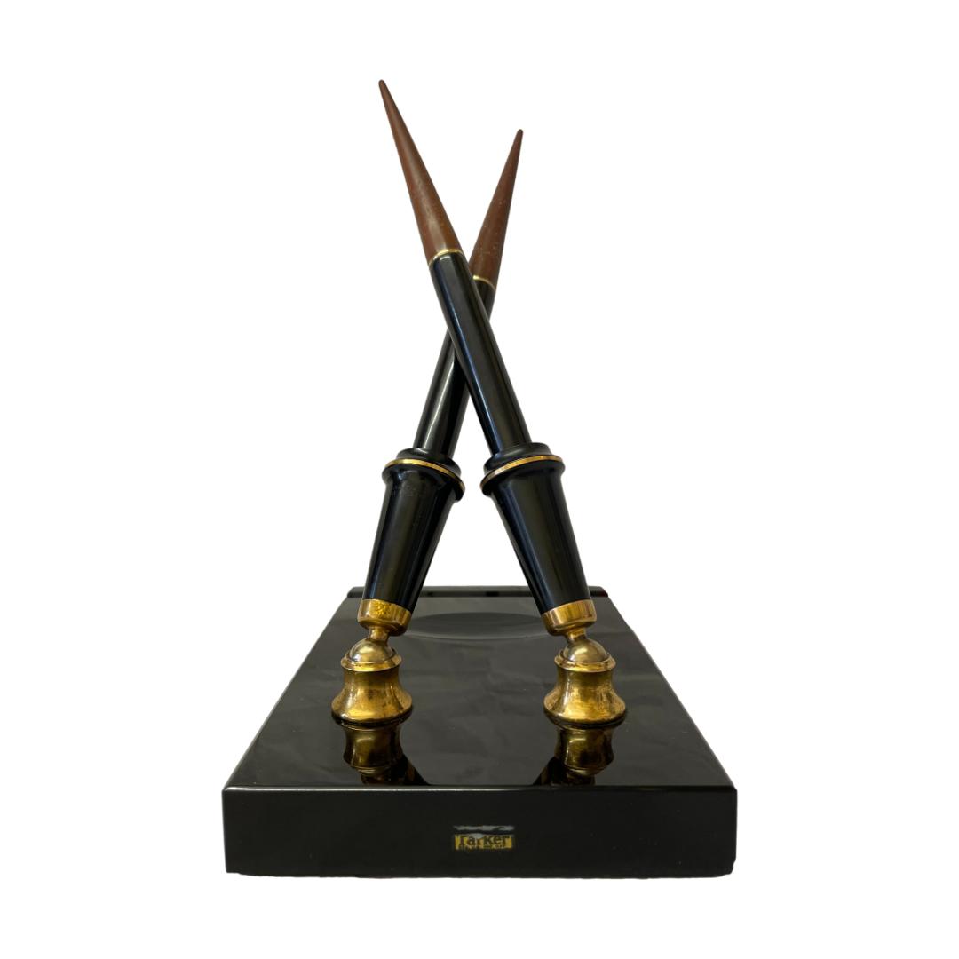 Vintage double desk set by Parker. Very chic desk top double pen set. The set consists of 2 Vacumatic fountain pens, one with 14kt gold fine point nib and other with a gilt metal italic nib. The stand, in smooth black granite like stone has a pen rest and an oval space probably for the ink bottle. The whole set is colour coordinated in black and brass with brown to the pen ends which twist off to reveal the filling mechanism. Each pen measures 210mm long. Pen holder slots are 75mm tall. Stand measures 75mm long, 113mm wide and 22mm. Main photo of the stand seen with the pen stand end in the foreground with the original worn Parker label visible in the foreground in yellow.