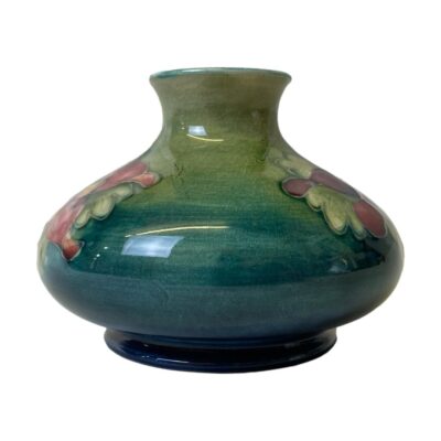 Vintage Moorcroft vase in orchid pattern. Small bulbous Moorcroft vase in sea green/blue with a pink and mauve orchid to either side. Moorcroft stamp to the base. In good condition with only minimal crazing. c1940s. Measures 68mm in diameter at base, 105mm at widest and 40mm across the top. Photo of vase from an eye level and showing the non decorated plain sea blue green colouring of the vase. The tips of the flowers can be seen to the extreme left and right. The darker shade of blue green is an the bottom of the vase and graduates to lighter shade further up.