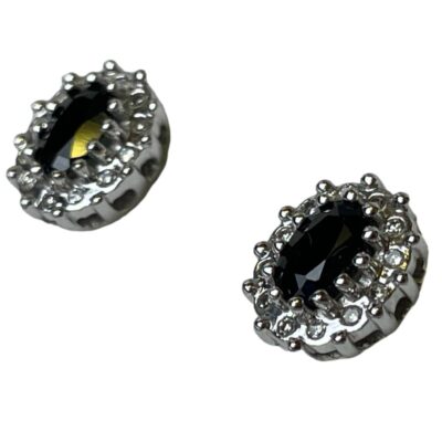 9 karat gold stud earrings with sapphire and diamonds. Pretty pair of 9 karat gold stud earrings with an oval cut sapphire to the centre framed by 12 small round cut diamonds. Hallmarked to the earring posts and butterfly fasteners. Earring front measure 10mm by 8mm. Photo of earrings seen from a diagonal side angle in close up with earring fronts facing upward towards the top left corner. The earring in the top left corner has a light reflection which is showing as a yellow dot - this is not part of the earring!