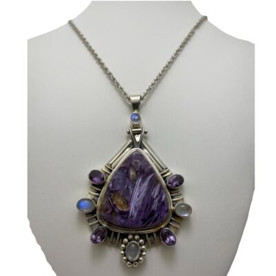 Statement charoite pendant on silver chain. Large striking sterling silver pendant with a substantial charoite stone to the centre and amethyst & rainbow moonstones evenly distributed around it and set in sterling silver. A rainbow moonstone also adorns the unusual bail. Hallmarked 925 to back of pendant and clasp. Pendant measures 88mm long from top of bail and 55mm at widest. Main photo of necklace displayed on a stand with the charoite pendant facing forward. The bail has a rainbow moonstone set on it then sterling silver frame around the triangular pyramid shaped charoite with alternating amethyst and moonstones set around the lower half of the triangle shaped stone.