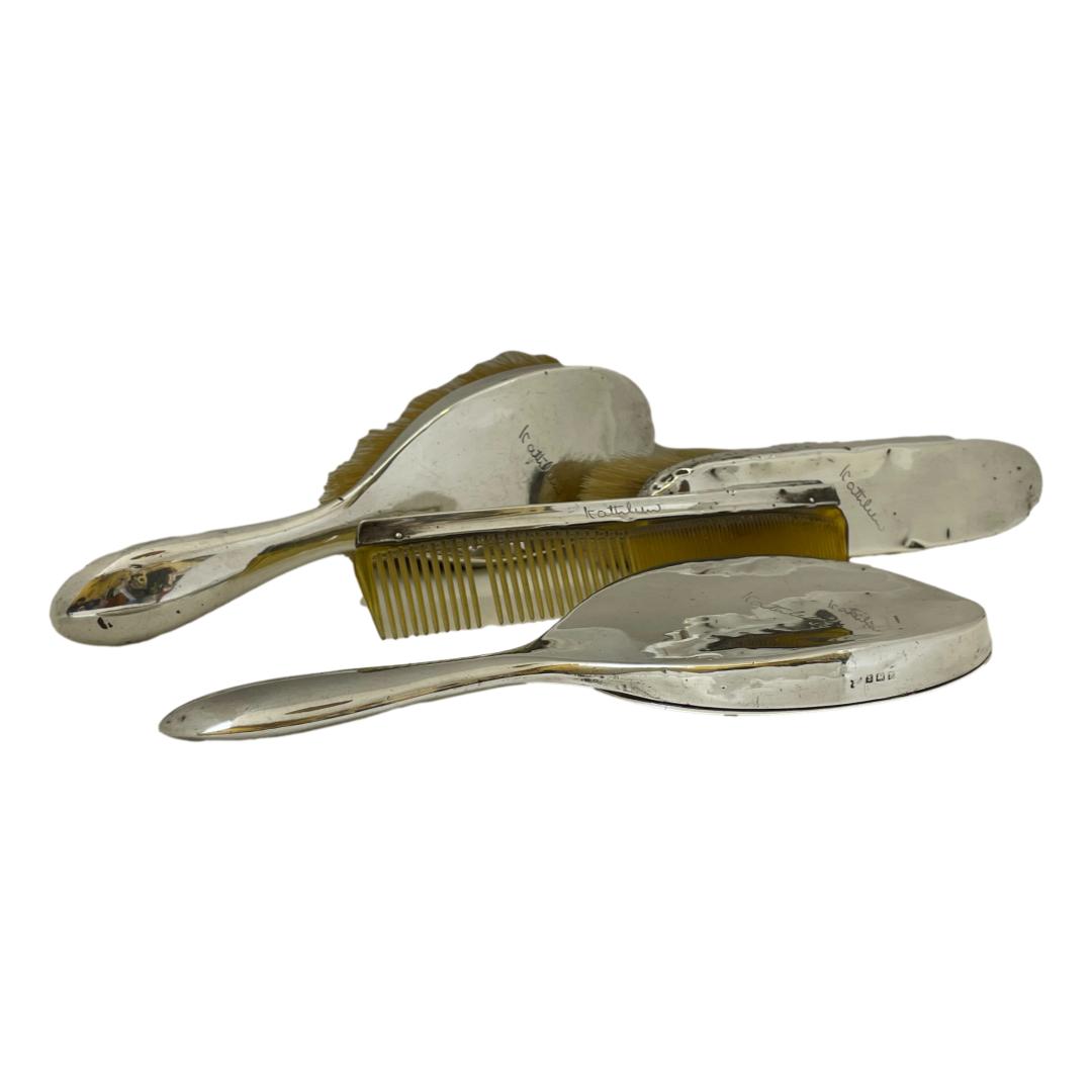 Antique sterling silver dressing table vanity set. c1923 Birmingham assayed sterling set comprising of a hand mirror, hairbrush, clothes brush and comb. All pieces in fair condition with minor dinks except for the clothes brush which has more dinks than others. The comb is missing a tooth and the silver backed mirror has some minor foxing nearer the handle. All pieces have been personalised to Kathleen. Comb measures 185mm by 40mm. Clothes brush is 200mm long at bristles, 40mm wide & 45mm deep. The hairbrush is 245mm long, 65mm at widest, 47mm deep. The mirror is 250mm long, 105mm wide and 16mm deep. Total weight is approximately 20ounces. Main photo of all 4 pieces displayed together. The handles of the hairbrush and mirror are on the left. The mirror is face down in the foreground with comb behind propped up against the back of the clothes brush and the hairbrush on its side in the background.
