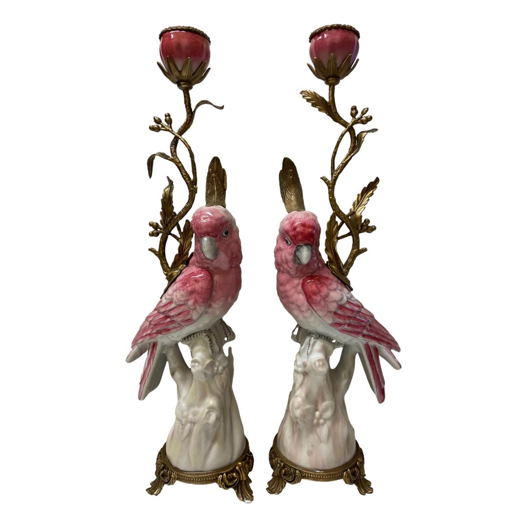 A pair of pink parrot candlesticks. An elegant pair of modern hand-painted porcelain parrots in pink with ormolu accents on a tripod base in the style of the sun king Louis XIV. Stamped to the base. The pair are not identical in size or colour which adds extra charm to this pretty pair. Approximate measurements 425mm tall, 135mm at widest and 82mm at deepest for one. Main photo of the 2 candlesticks displayed side by side with the birds facing forward towards camera. The camera angle is slightly raised but shows the whole of each candlestick. The birds are pink as are the sconces and the base is in white with gold ormolu at the base, birds head feathers and branch that comes out of the birds backs which holds the sconces. From the front view, the branches appear behind the birds.