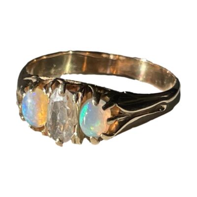 Victorian ring with diamond and opals. Stunning antique ring set with an old cut oval diamond to the centre with an opal cabochon to either side. The opals have lovely colour play with one mainly in green and other blue with flashes of pink, orange & yellow. The ring is unmarked and has been tested as 18 karat gold. Size N / 6.75. Weight 3.2grams. Photo of ring on a flat surface with the front facing towards the light on the left. The lefthand opal is flashing blue and peach and yellow and righthand one is more green blue and peach.