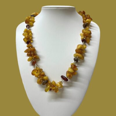 Main photo showing necklace displayed on a stand and seen looking straight on from the front. The necklace is generous in length and hangs below the collar bone. The background colour is a dark mustard which enhances the colours of the amber pieces.