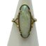 Main photo of ring displayed on a cone shaped stand. The ring front is forward facing. The opal is of a long oval shape and set on split band shoulders. The opal is a milky white with flashes of good colour. In this photo, there are flecks of yellow, green, lilac and pink.