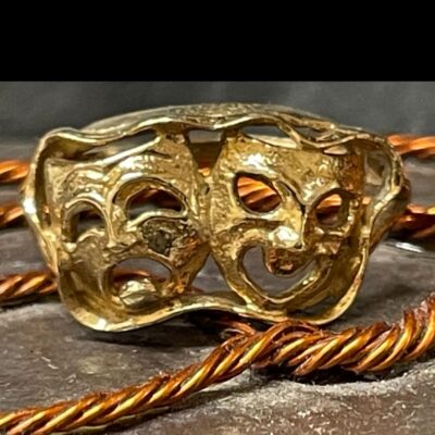 Main photo of ring displayed against a background of copper twists on stone. The ring is seen from the front with the tragedy mask on the left and comedy to its right. The eyes and mouth of both masks are open through. A lovely gift for someone who is wild about the theatre!