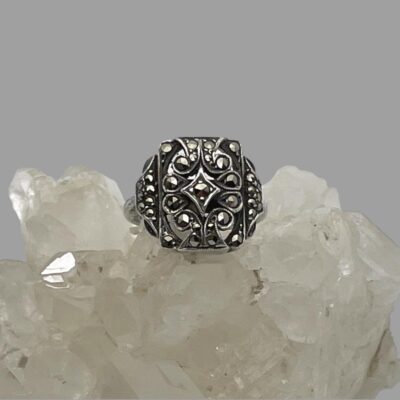 Main photo of ring displayed on top of some quartz crystal and seen with ring front facing forward. The marcasite pieces are catching the light and sparkling nicely. The design of the front is very Art Deco. A diamond shape at the centre holds one marcasite. The overall shape of the front is rectangular but without sharp edges .