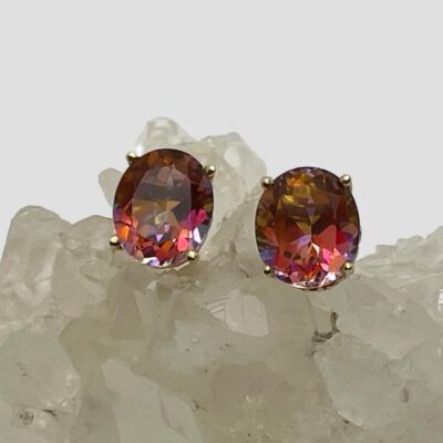 Main photo of earrings displayed on top of a cluster of quartz crystal with the earrings fronts forward facing. The azotic mystic topaz stones are beautifully facet cut ovals with an array of different colours in an assortment of pink hues.