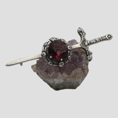 Main photo of brooch displayed on top of a small lump of natural amethyst of a lilac colour which contrasts to the darker purple colour of the amethyst on the brooch. The sword hilt is to the right.