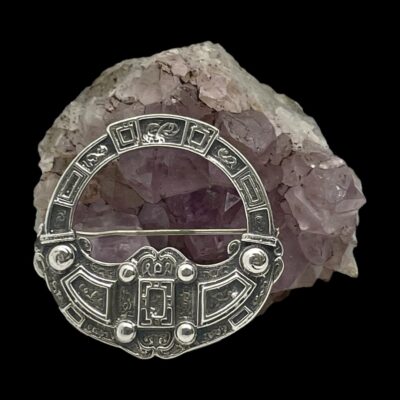 Main photo of brooch shown leaning up against a cluster of natural amethyst with the brooch front forward facing. The safety chain and pin is hidden behind the brooch so only the brooch front is visible with the Celtic signs clearly on display.