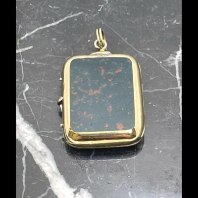 Photo of locket with the bloodstone side on display, the bail to the top and opening side on the left and displayed on a slab of black marble with white line inclusions. The bloodstone is a dark green with flecks of red hence its name.