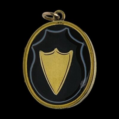 Photo of locket seen from the front and set against black background. The banded agate has been expertly cut with the white band within the agate complimenting and framing the golden shield at the centre.