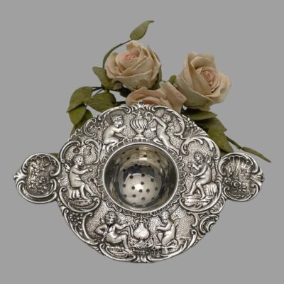 Main photo of strainer seen leaning against a small arrangement of pink roses. The strainer is shown woth the thumb rests to the left and right and the cherubs facing the right way up. The strainer is in circular form with the thumb rests jutting out. The 2 cherubs at the bottom are tending the fire for boiling the kettle. The cherub on the left and right thumb rests are pouring milk and drinking tea (one on left looks like it is drinking tea from saucer!) and the 2 cherubs at the top are tending to the tea pot. A really lovely item for making the perfect cup of tea!