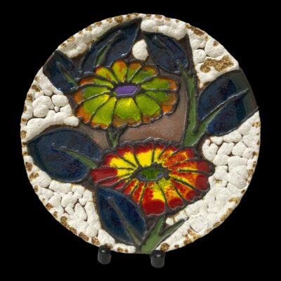 Main photo of plate displayed on a plate stand (2 dark wood feet visible at bottom of plate) The plate has a large glossy green and red flower to the centre with blue/green foliage around them. The outer area of the plate has white lava which looks a little like popcorn or rice cakes.