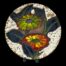 Main photo of plate displayed on a plate stand (2 dark wood feet visible at bottom of plate) The plate has a large glossy green and red flower to the centre with blue/green foliage around them. The outer area of the plate has white lava which looks a little like popcorn or rice cakes.