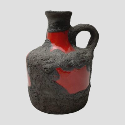 Main photo of jug seen from an eye level with the single handle to the right. The jug is mostly covered with black lava with a few small windows of gloss red which creates a striking contrast. This side of the jug has a small red gloss window on the neck and a larger one just below the handle.