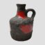 Main photo of jug seen from an eye level with the single handle to the right. The jug is mostly covered with black lava with a few small windows of gloss red which creates a striking contrast. This side of the jug has a small red gloss window on the neck and a larger one just below the handle.